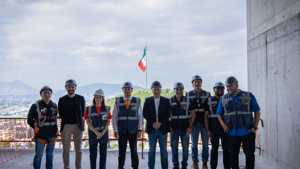 Group of Construction Workers and Engineers in Hard Hats and Safety Vests Pose on a Rooftop Overlooking a City, with an Italian Flag in the Background. - Tu Vecino Informa