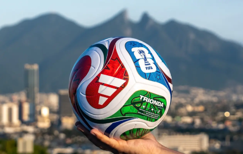 Image - Tu Vecino Informa Close-up of a Multicolored Fifa Soccer Ball Held by a Hand with a City and Mountains in the Background. - Tu Vecino Informa