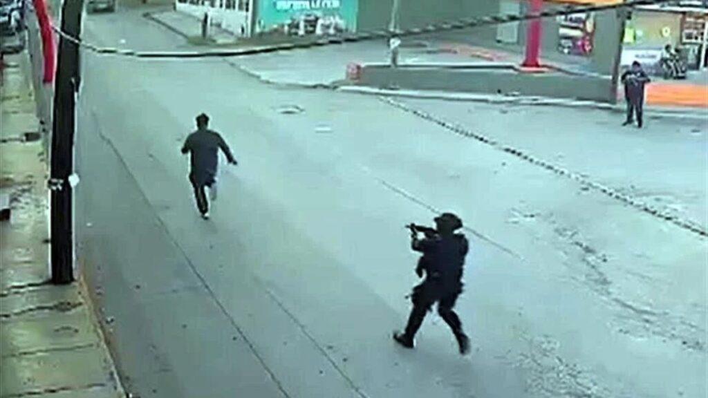 a Man in Dark Clothing Runs Down a Wet City Street While a Gun-wielding Figure Pursues Him from Behind, Camera-style Surveillance View. - Tu Vecino Informa