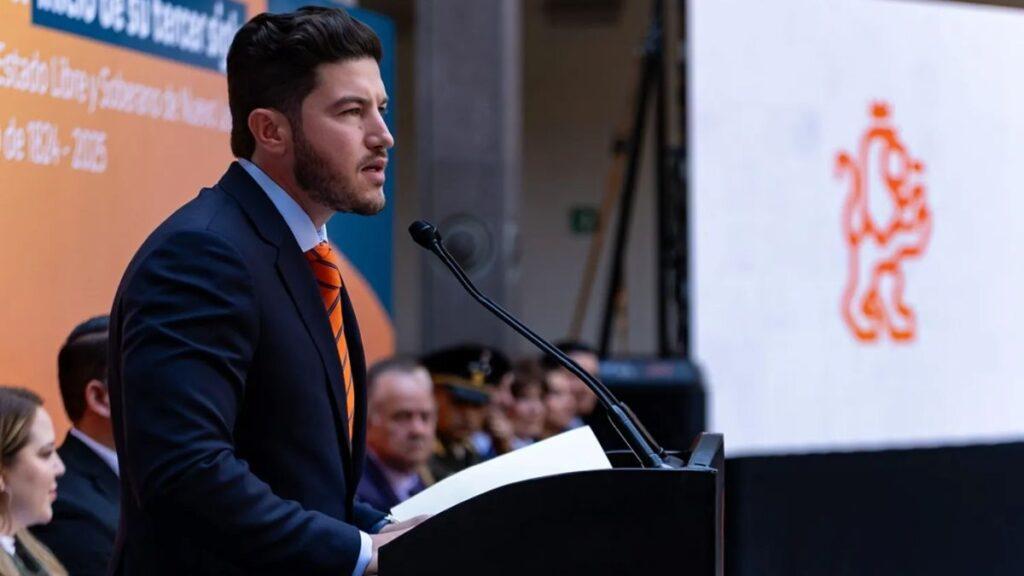 Samuel-garcía-sat-megadeuda-nuevo-león-sat-acorrala-a-samuel - Tu Vecino Informa Man in a Dark Suit and Orange Striped Tie Speaks at a Podium During an Outdoor Ceremony, with a Large Orange Crest on a Screen Behind Him. - Tu Vecino Informa