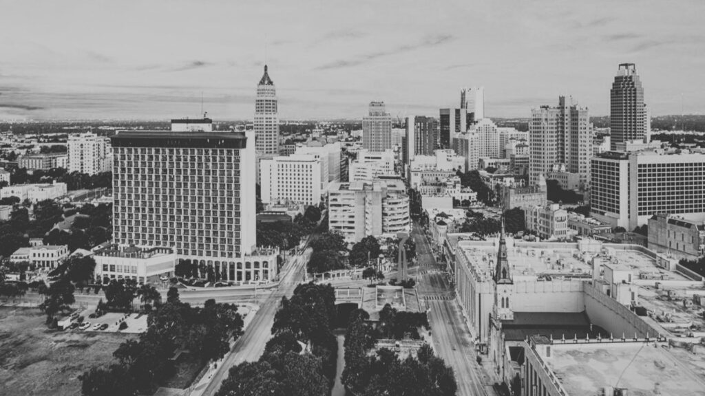 Gira-de-trabajo-en-san-antonio 1 - Tu Vecino Informa Aerial View of a Downtown City Skyline in Grayscale, with Tall Skyscrapers and a Wide Avenue Cutting Through the Center. - Tu Vecino Informa