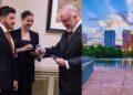 Three Professionals in Suits Exchange an Award Indoors, Smiling Near a Framed Picture on the Wall (left Side). the Right Side Shows a Modern City Skyline by a River with a Wooden Boardwalk. - Tu Vecino Informa