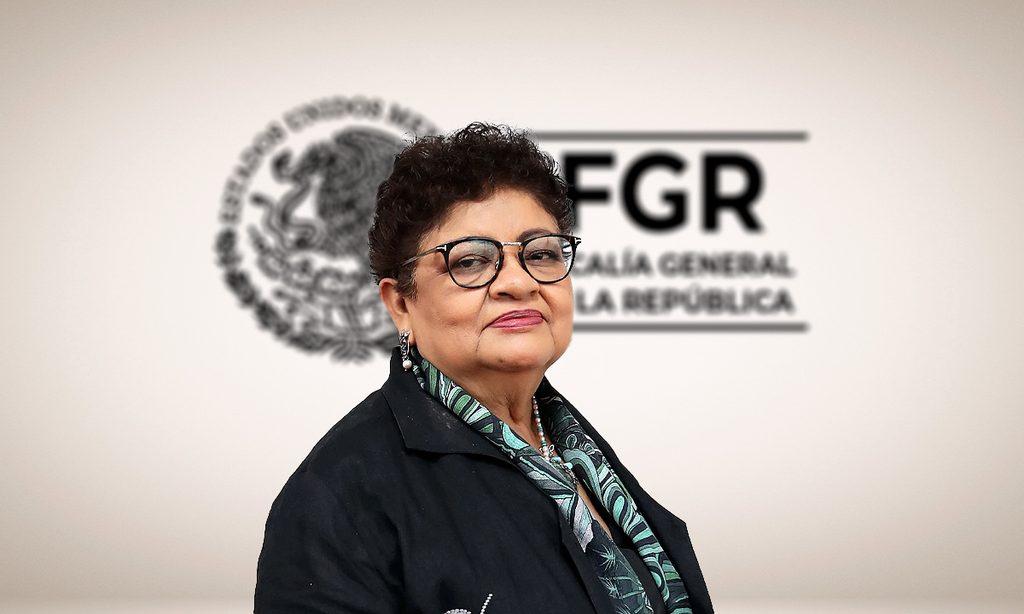 Ernestina_godoy_fgr-1 - Tu Vecino Informa Portrait of a Middle-aged Woman with Short Curly Hair and Glasses, Wearing a Black Blazer and Green Scarf, in Front of a Logo Backdrop. - Tu Vecino Informa