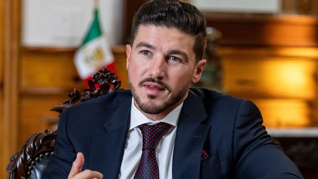 Tu-vecino-informa-gobernador-nuevo-leon-publicidadjpg - Tu Vecino Informa Man in a Navy Suit and Red Tie Speaks, Gesturing with His Hand in a Wood-paneled Office Setting with a Flag in the Background. - Tu Vecino Informa
