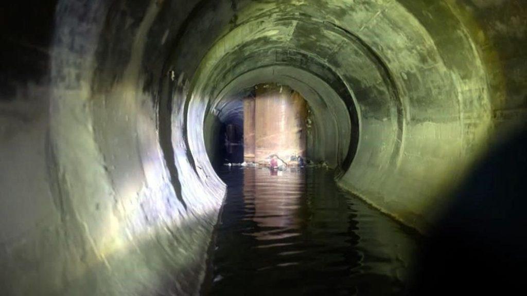 Long Concrete Tunnel with Standing Water, View Toward a Distant Circular Opening; a Person Appears Near the Far End. - Tu Vecino Informa